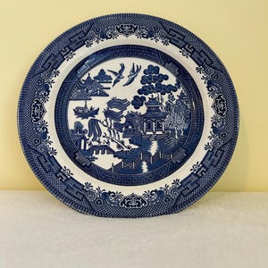 May include: A round, blue and white Willow pattern plate. The plate features a detailed landscape scene with pagodas, trees, and a boat. The rim has a decorative border. This vintage-style plate is suitable for serving food or as a decorative piece.