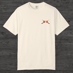 Puede incluir: Camiseta color crema con un diseño bordado de una bola de billar y tacos cruzados. El diseño incluye una bola 8 amarilla y una bola 9 roja. La camiseta tiene mangas cortas y cuello redondo.