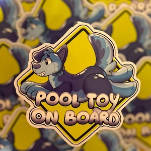 May include: A sticker featuring a cartoon canine character in shades of blue and purple. The character is lying down with a smiling expression. The sticker has the text "POOL TOY ON BOARD" in a white, outlined font, set against a yellow diamond.