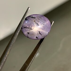 May include: An oval-shaped, translucent light purple gemstone with a cabochon cut. The stone features a distinct six-rayed star effect centred on its surface, accompanied by several dark, needle-like inclusions scattered throughout the interior.