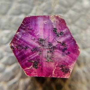 May include: A hexagonal raw ruby crystal featuring a vibrant magenta and deep pink color palette. The surface displays a translucent quality with visible internal striations and scattered dark black mineral inclusions across the flat face.