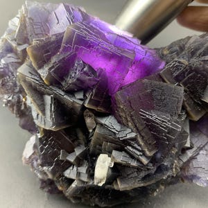 Puede incluir: Primer plano de un espécimen mineral de fluorita. La fluorita presenta un tono púrpura intenso en el centro, que se transforma en tonos más oscuros de gris y negro en los bordes. La estructura cristalina es visible, con formaciones geométricas nítidas.