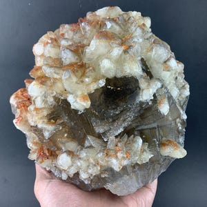 Puede incluir: Una gran formación de cristal en bruto. El cristal presenta secciones claras, blancas y gris ahumado, con acentos de color naranja y marrón rojizo. Los bordes naturales ásperos y las texturas variadas crean un espécimen mineral único.