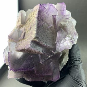 Puede incluir: Un gran grupo de cristales de fluorita natural. El cristal presenta una mezcla de formaciones cúbicas moradas y claras, con algunas áreas que muestran un color beige claro. La estructura general es compleja y visualmente impactante, mostrando la belleza natural del mineral.