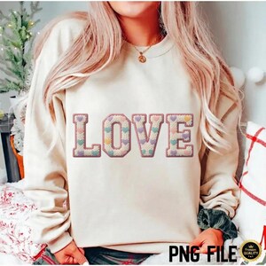 Puede incluir: Una sudadera color crema con la palabra "LOVE" bordada en la parte delantera. Las letras están rellenas de pequeños corazones en colores pastel y delineadas en un rosa más oscuro. Las mangas son largas y el estilo es informal.