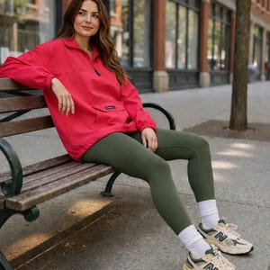 Peut inclure: Une veste pull rouge vif à manches longues avec un col zippé et une poche avant, associée à un legging vert olive uni, des chaussettes blanches et des baskets de sport beiges et vertes avec la lettre N sur le côté.