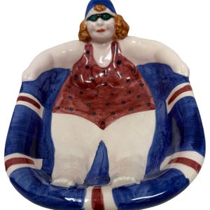 May include: A ceramic figurine of a woman in a red polka-dot swimsuit and blue swim cap, wearing sunglasses. The figurine is seated in a blue dish with red and white stripes, resembling a Union Jack flag.