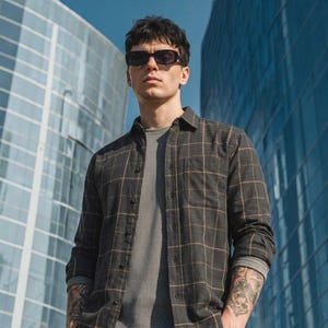 May include: A dark gray button-down shirt with a thin tan grid pattern worn over a gray thermal long-sleeve shirt. The shirt features a single chest pocket and is paired with black sunglasses. The outfit is worn against a backdrop of glass buildings.