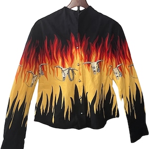 Puede incluir: Una camisa abotonada de manga larga con un llamativo diseño de llamas en rojo, naranja y amarillo. La camisa presenta un cuello y puños negros, y un patrón repetido de calaveras de toro con detalles de cuerda. La camisa está en una percha de madera.