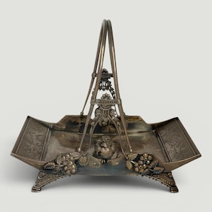 May include: A silver-toned metal basket with a high arched handle, featuring intricate embossed floral and berry motifs along the sides. A small sculpted bird figure sits at the base of the handle, and the basket rests on four decorative feet.
