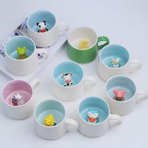 May include: A selection of ceramic mugs featuring animal figurines. The mugs are white with coloured interiors, including blue, green, and pink. Each mug has a different animal, such as a panda, frog, cow, and pig. The mugs have a handle.