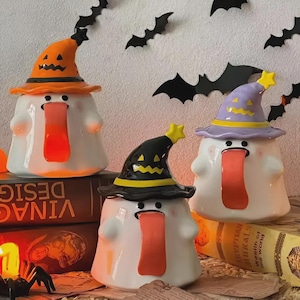 May include: Three white ceramic ghost figurines wearing orange, black, and purple witch hats. Each ghost has a long, red tongue. Black bat decorations are on the wall. The figurines are Halloween-themed and decorative.