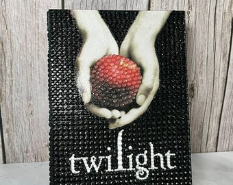 May include: A black book cover with the title "twilight" in white lettering. The cover features a graphic of two hands holding a red apple. The author's name, Stephenie Meyer, is printed below the title.
