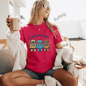 Puede incluir: Una camiseta magenta con el texto "BINARY BUDDIES" y tres robots de dibujos animados. Los robots son amarillos, azules y naranjas, cada uno con un número binario en el pecho. La camiseta está siendo usada.