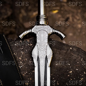 May include: A detailed, silver-colored sword with an ornate handle and a black blade, resting on a wooden surface. The handle features intricate designs and a crossguard. A black sheath is visible to the left of the sword.