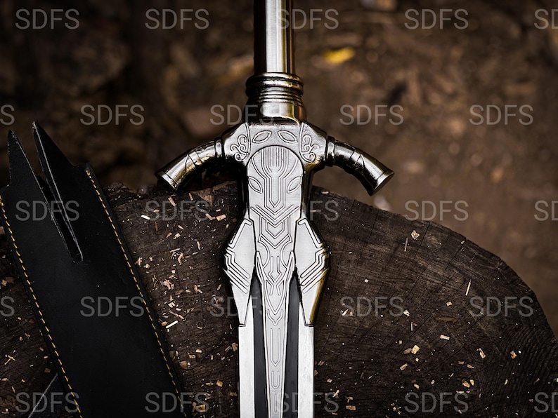 May include: A detailed, silver-colored sword with an ornate handle and a black blade, resting on a wooden surface. The handle features intricate designs and a crossguard. A black sheath is visible to the left of the sword.