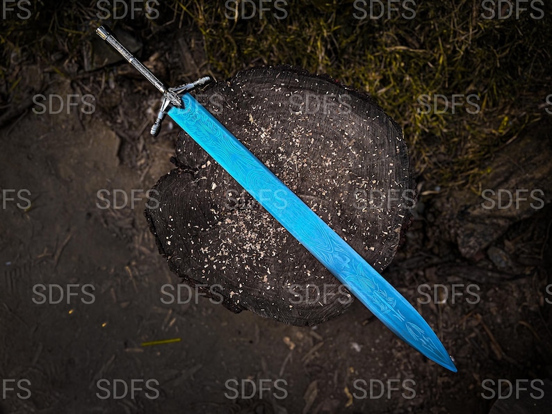 May include: A blue sword with a detailed blade design rests on a dark wooden surface. The sword has a silver-colored hilt and crossguard. The background features dark earth and foliage, creating a contrast with the bright blue blade.