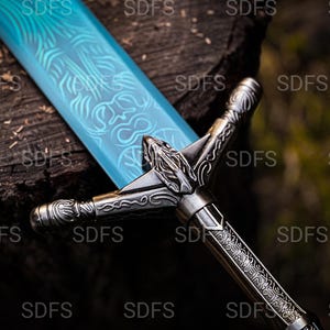May include: A detailed image of a fantasy sword with a light blue blade featuring intricate etched designs. The hilt and crossguard are silver-toned with ornate carvings. The sword rests on a dark, textured wooden surface.