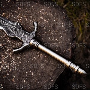 May include: A detailed, silver-colored sword with an ornate handle and blade design. The sword is resting on a weathered, dark brown wooden surface, with wood shavings scattered around it. The sword's blade is approximately 24 inches long.