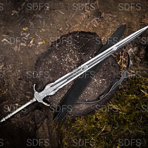 May include: A silver and black sword with a decorative handle and a long, pointed blade rests on a tree stump. A black sheath with a shoulder strap lies beside the sword. The background is a mix of wood and foliage.