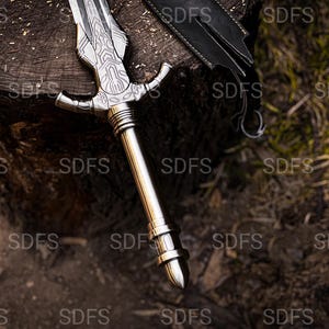 May include: A silver-colored sword with an ornate handle and blade rests on a wooden surface. The blade features detailed engravings, and the handle has a cross-guard design. A black leather sheath is partially visible.