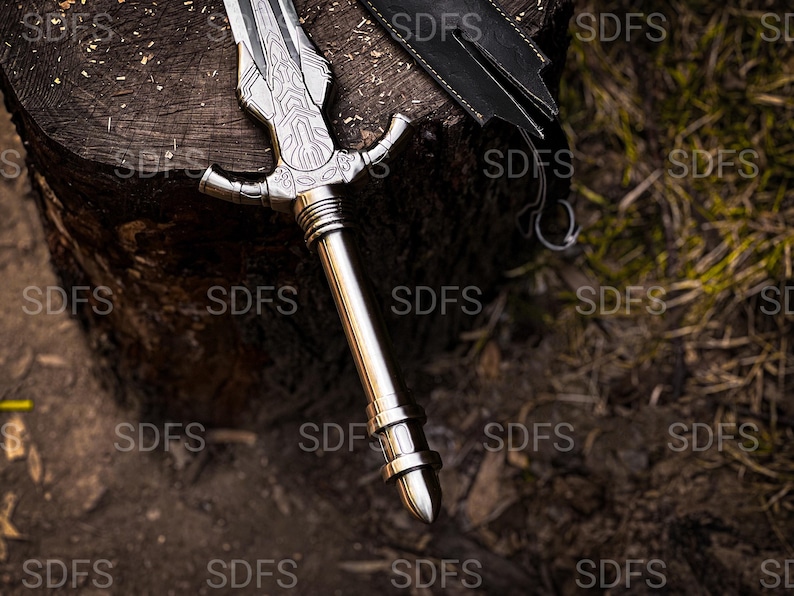 May include: A silver-colored sword with an ornate handle and blade rests on a wooden surface. The blade features detailed engravings, and the handle has a cross-guard design. A black leather sheath is partially visible.
