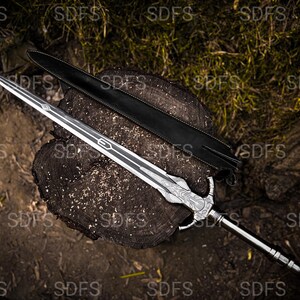 May include: A detailed image of a silver sword with a decorative handle and a black leather sheath. The sword is resting on a wooden surface, showcasing its intricate design and craftsmanship. The blade has a length of approximately 30 inches.