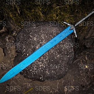 May include: A blue sword with a detailed pattern on the blade rests on a dark wooden surface. The sword has a silver hilt and a pointed tip. The background is a mix of wood and foliage.