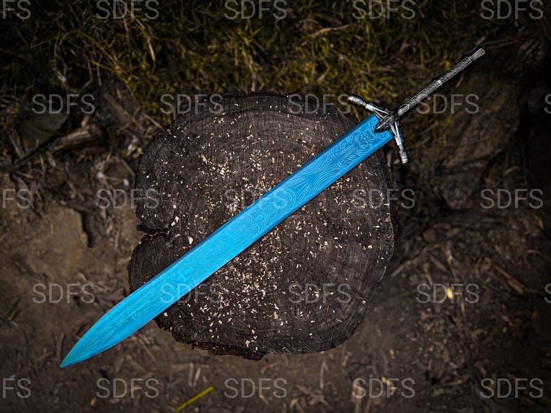 May include: A blue sword with a detailed pattern on the blade rests on a dark wooden surface. The sword has a silver hilt and a pointed tip. The background is a mix of wood and foliage.