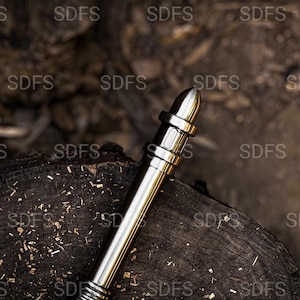 May include: A metallic silver pen with a bullet-shaped tip rests on a dark wooden surface. The pen has a cylindrical body with several bands and a textured grip. The background is a blurred view of wood and natural elements.