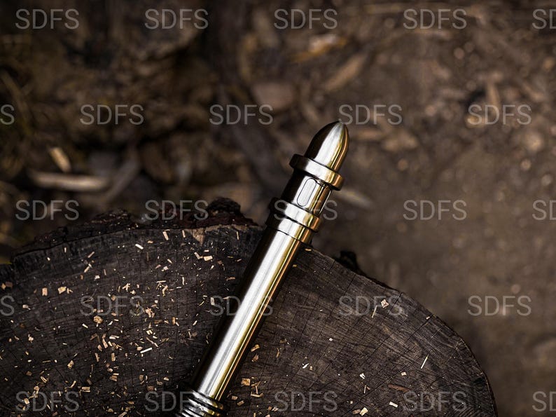 May include: A metallic silver pen with a bullet-shaped tip rests on a dark wooden surface. The pen has a cylindrical body with several bands and a textured grip. The background is a blurred view of wood and natural elements.
