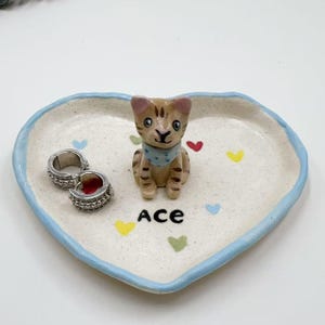 Op de afbeelding: Hartvormige keramische sieradenschaal met een lichtblauwe rand. De schaal heeft een kleine, bruine kattenfiguur met een blauwe bandana en het woord "Ace". Twee zilveren oorringen liggen aan de linkerkant van de schaal.