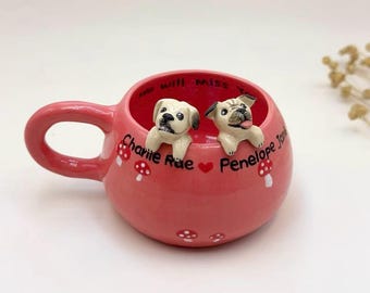 Puede incluir: Una taza de cerámica rosa con asa, con dos pequeñas figuras de perros en el interior. La taza está decorada con diseños de setas y los nombres "Charlie Rae" y "Penelope Jo" están escritos en el lateral.
