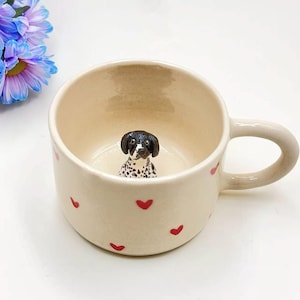 May include: Cream ceramic mug decorated with red hearts. Inside the mug is a small dog figurine with black and white spots. The mug has a curved handle. The background is white.