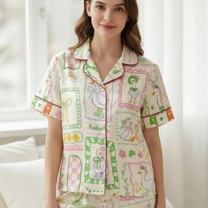 Puede incluir: Conjunto de pijama color crema con un estampado caprichoso de gansos. La camiseta de manga corta y los pantalones cortos presentan un patr&oacute;n repetido de gansos enmarcados, lazos y otros elementos decorativos en rosa, verde y morado. El conjunto est&aacute; ribeteado con un ribete burdeos.