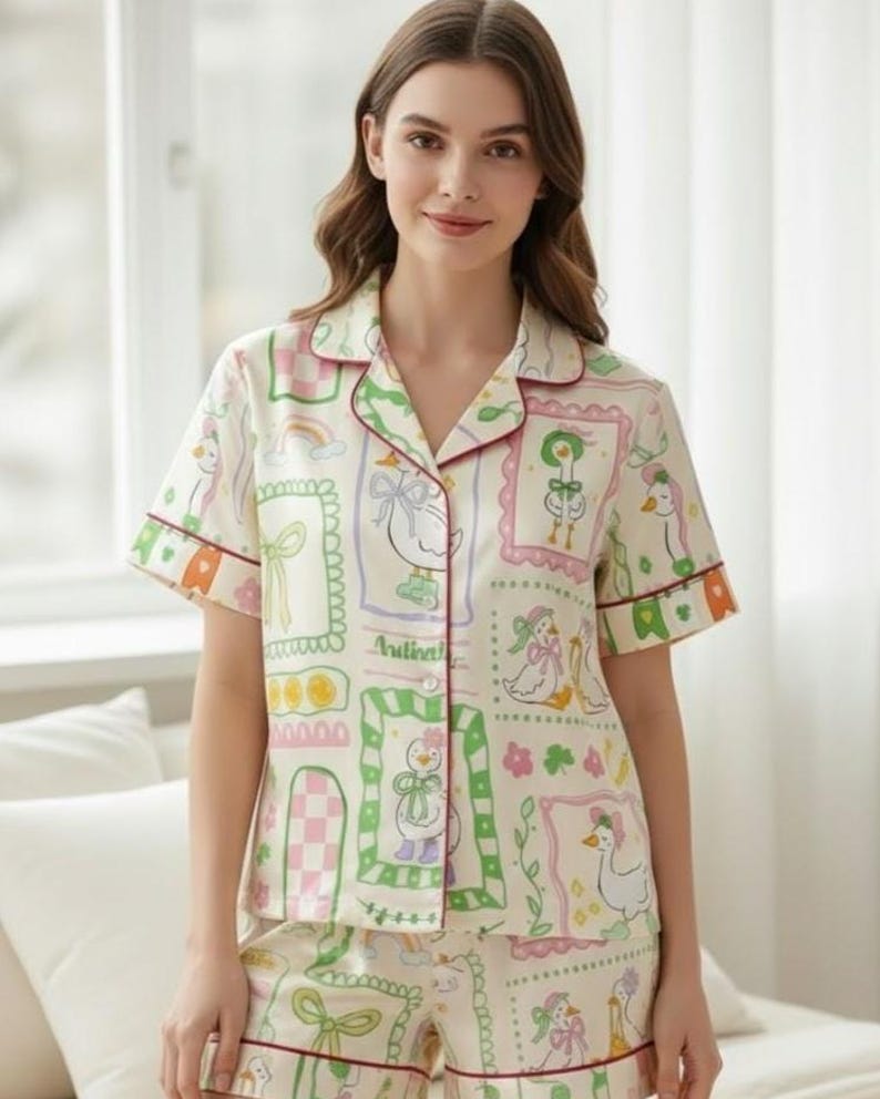 Puede incluir: Conjunto de pijama color crema con un estampado caprichoso de gansos. La camiseta de manga corta y los pantalones cortos presentan un patr&oacute;n repetido de gansos enmarcados, lazos y otros elementos decorativos en rosa, verde y morado. El conjunto est&aacute; ribeteado con un ribete burdeos.