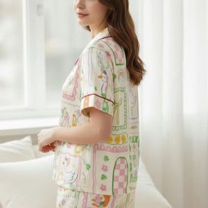 Puede incluir: Conjunto de pijama color crema con un estampado caprichoso de patos, arco&iacute;ris y elementos florales. La parte superior de manga corta y los pantalones cortos est&aacute;n ribeteados con un ribete burdeos. El conjunto presenta una camisa con botones y pantalones cortos con cintura el&aacute;stica.