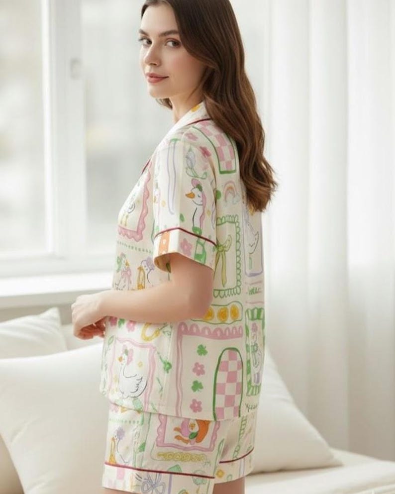 Puede incluir: Conjunto de pijama color crema con un estampado caprichoso de patos, arco&iacute;ris y elementos florales. La parte superior de manga corta y los pantalones cortos est&aacute;n ribeteados con un ribete burdeos. El conjunto presenta una camisa con botones y pantalones cortos con cintura el&aacute;stica.