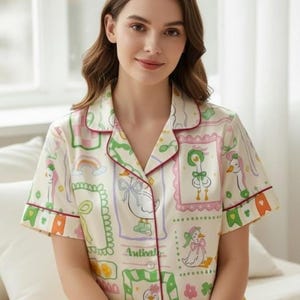 Puede incluir: Conjunto de pijama color crema con un estampado caprichoso de patos. La parte superior de manga corta y los pantalones cortos presentan un dise&ntilde;o repetido de patos en marcos, arco&iacute;ris y detalles florales, delineados en rojo y verde. El conjunto tiene un ajuste relajado.