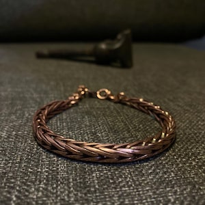 K&ouml;nnte beinhalten: Ein kupferfarbenes Armband mit einem geflochtenen Kettenmuster. Das Armband besteht aus kleinen, ineinandergreifenden Gliedern, die eine strukturierte Oberfl&auml;che bilden. Der Verschluss ist sichtbar, und das Armband liegt auf einer strukturierten Oberfl&auml;che.