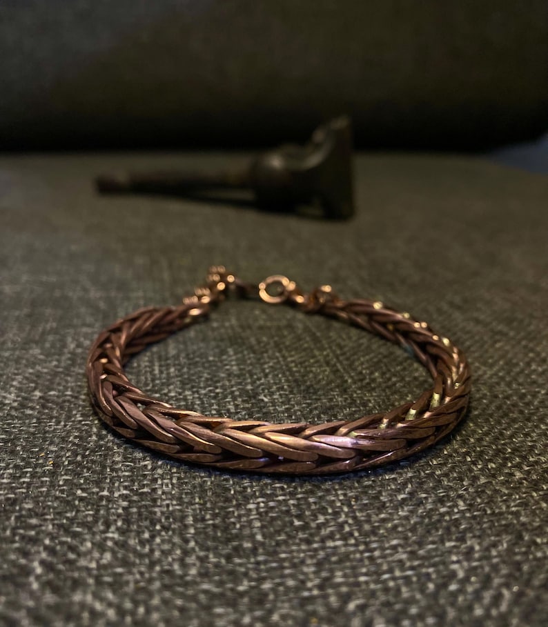 K&ouml;nnte beinhalten: Ein kupferfarbenes Armband mit einem geflochtenen Kettenmuster. Das Armband besteht aus kleinen, ineinandergreifenden Gliedern, die eine strukturierte Oberfl&auml;che bilden. Der Verschluss ist sichtbar, und das Armband liegt auf einer strukturierten Oberfl&auml;che.
