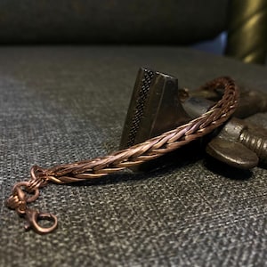 K&ouml;nnte beinhalten: Ein kupferfarbenes geflochtenes Armband mit einem Karabinerverschluss. Das Armband besteht aus ineinander verwobenen Metallstr&auml;ngen, wodurch ein strukturiertes, gewebtes Aussehen entsteht. Das Armband wird auf einer dunkelgrauen Oberfl&auml;che pr&auml;sentiert.