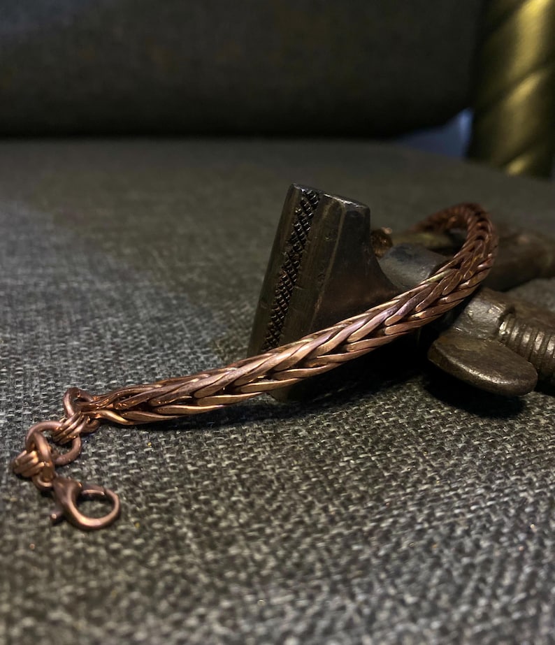 K&ouml;nnte beinhalten: Ein kupferfarbenes geflochtenes Armband mit einem Karabinerverschluss. Das Armband besteht aus ineinander verwobenen Metallstr&auml;ngen, wodurch ein strukturiertes, gewebtes Aussehen entsteht. Das Armband wird auf einer dunkelgrauen Oberfl&auml;che pr&auml;sentiert.