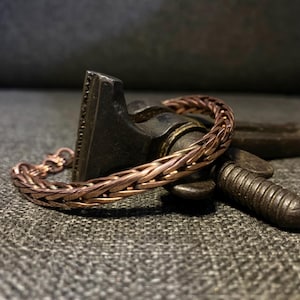 K&ouml;nnte beinhalten: Ein kupferfarbenes geflochtenes Armband mit einem Verschluss. Das Armband besteht aus miteinander verflochtenen Kupferdrahtstr&auml;ngen, wodurch ein strukturiertes, gewebtes Aussehen entsteht. Das Armband liegt auf einer dunklen, strukturierten Oberfl&auml;che.