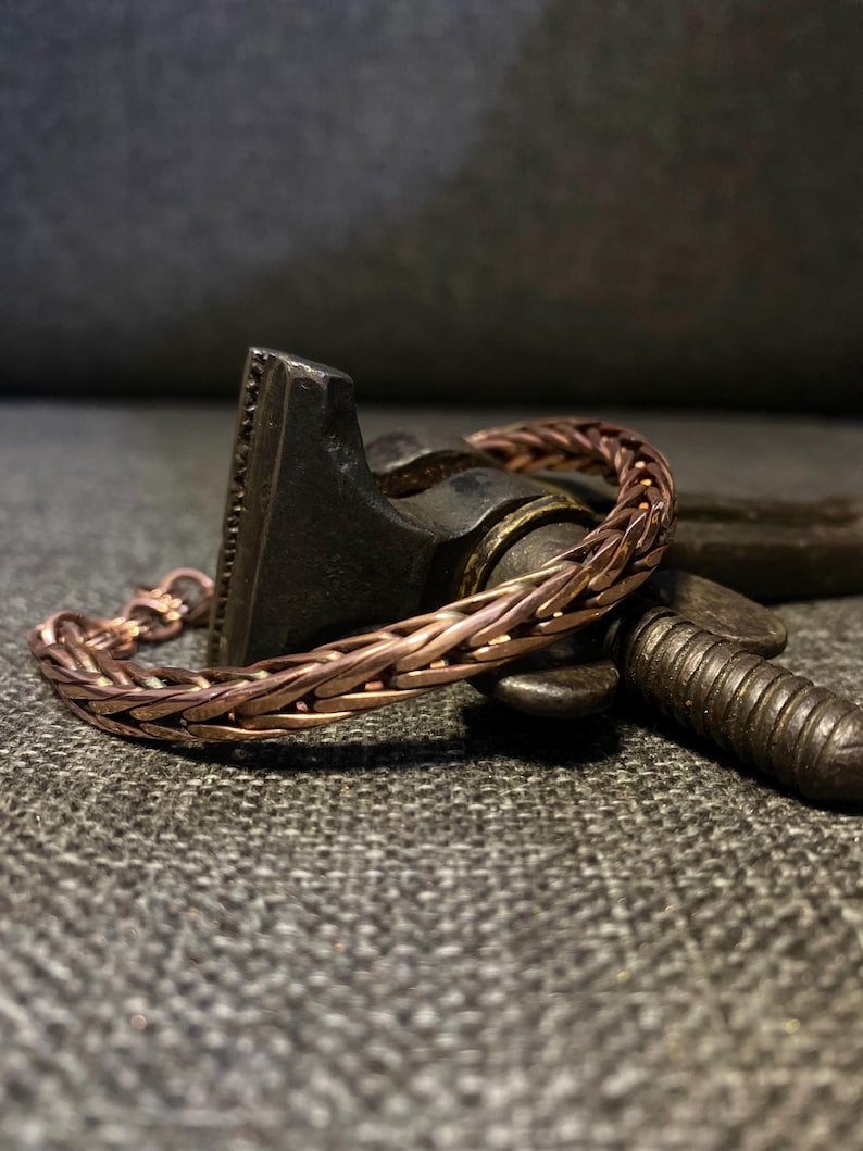 K&ouml;nnte beinhalten: Ein kupferfarbenes geflochtenes Armband mit einem Verschluss. Das Armband besteht aus miteinander verflochtenen Kupferdrahtstr&auml;ngen, wodurch ein strukturiertes, gewebtes Aussehen entsteht. Das Armband liegt auf einer dunklen, strukturierten Oberfl&auml;che.