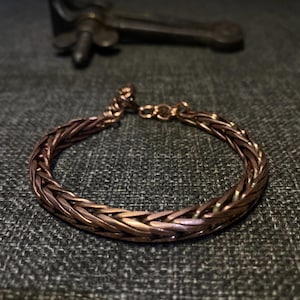 K&ouml;nnte beinhalten: Ein kupferfarbenes Armband mit geflochtenem Design. Das Armband ist kreisf&ouml;rmig und scheint aus mehreren Str&auml;ngen Kupferdraht zu bestehen. Im Hintergrund befindet sich ein dunkles Metallobjekt.