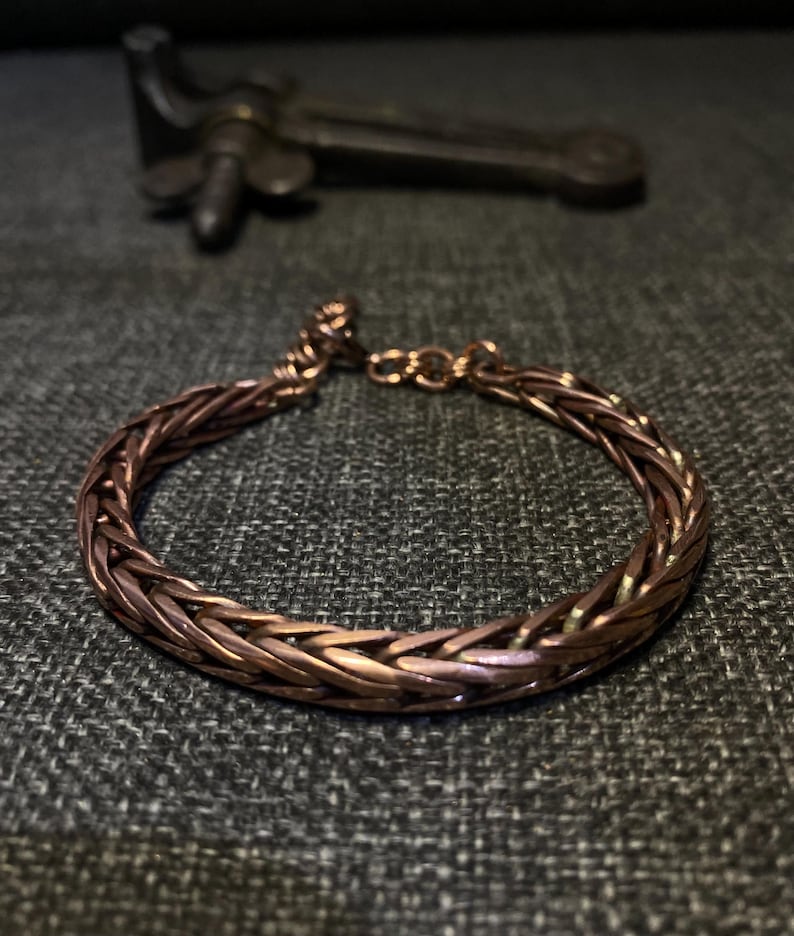 K&ouml;nnte beinhalten: Ein kupferfarbenes Armband mit geflochtenem Design. Das Armband ist kreisf&ouml;rmig und scheint aus mehreren Str&auml;ngen Kupferdraht zu bestehen. Im Hintergrund befindet sich ein dunkles Metallobjekt.