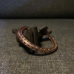 K&ouml;nnte beinhalten: Ein kupferfarbenes geflochtenes Armband. Das Armband besteht aus ineinander verschlungenen Metallstr&auml;ngen, wodurch ein strukturiertes, gewebtes Aussehen entsteht. Das Armband liegt auf einer dunklen, strukturierten Oberfl&auml;che.