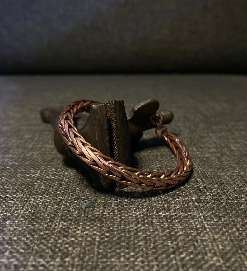 K&ouml;nnte beinhalten: Ein kupferfarbenes geflochtenes Armband. Das Armband besteht aus ineinander verschlungenen Metallstr&auml;ngen, wodurch ein strukturiertes, gewebtes Aussehen entsteht. Das Armband liegt auf einer dunklen, strukturierten Oberfl&auml;che.