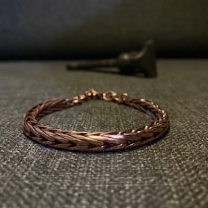 K&ouml;nnte beinhalten: Ein kupferfarbenes geflochtenes Armband mit strukturiertem Design. Das Armband besteht aus ineinandergreifenden Metallgliedern und hat einen Verschluss. Der Hintergrund ist eine dunkelgraue Oberfl&auml;che.